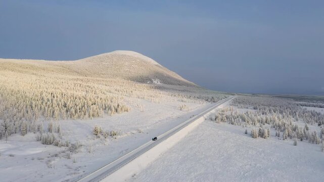 Aerial view of a lone car driving on a snowy road cutting through an expansive, snow-laden landscape under a muted sky, Oymyakon, Sakha Republic, Russia.