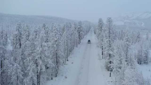 Aerial view of a lone car driving on a straight snowy road surrounded by snow-covered trees, Oymyakon, Sakha Republic, Russia.