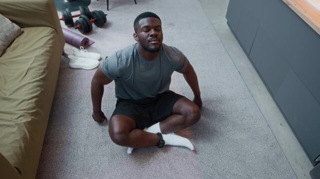 High angle shot of young African American man under house arrest sitting on floor in lotus position, taking deep breath and looking up, relaxing after yoga practice
