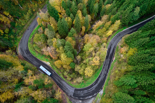 Aerial view of a winding road cutting through a vibrant forest with a lone blue bus navigating the curve, Oltre il Colle, Lombardy, Italy.