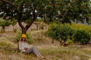 A young Caucasian woman with long hair sits under a peach tree in a grassy field. She wears a yellow hat and relaxed clothing, enjoying a peaceful moment in nature.
