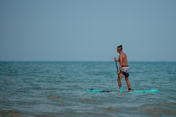 A young Caucasian man with short brown hair stands on a paddleboard in the ocean. He wears black...