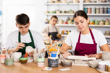 Concentrated young man and woman in aprons making handmade cups of row clay material in pottery workshop