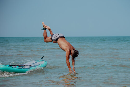 A young Caucasian man jumps into the ocean from a surfboard. The scene captures a sunny day at the beach with clear blue water.