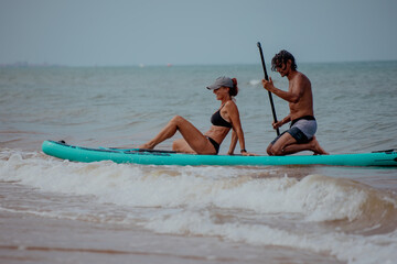 A young Caucasian man and a young Hispanic woman enjoy paddleboarding in the ocean. The man holds a...
