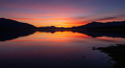 Naklejka premium Serene sunset reflecting on a calm lake, silhouetted mountains at dusk.