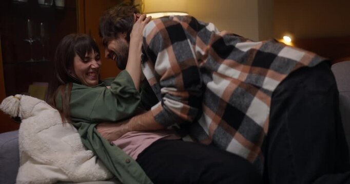 Cheerful guy plays with his girlfriend and gently squeezes her during cozy fun relationship of romantic couple on sofa in cozy apartment at home