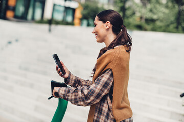 Young smiling woman using smartphone alongside an electric scooter in a modern urban environment during a sunny day
