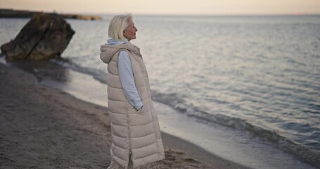 Mature woman with white hair in jacket walking along sea on beach enjoying sea view and fresh air during her walk during daytime