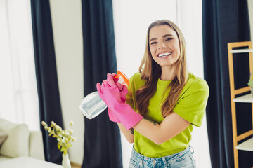 Smiling young woman in a living room wearing pink gloves and holding a spray bottle, promoting...