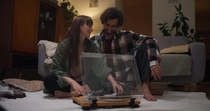 Happy girl and guy sitting near vinyl player and listening to music on record during cozy music and vintage style evening at home in cozy apartment in evening