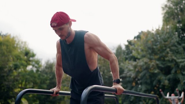A determined male athlete performs dips on parallel bars in an outdoor park setting. He focuses on his fitness routine, showcasing strength and dedication.park backdrop adds to the tranquil atmosphere