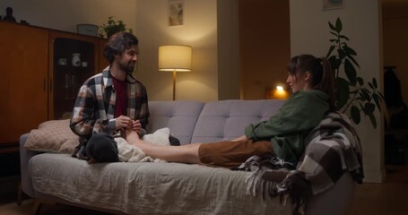 Happy brunette guy gives gentle foot massage to his girlfriend during rest and romantic conversation in cozy apartment at home