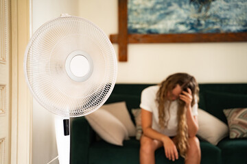 Woman sitting on sofa feeling unwell from summer heatwave, electric fan in foreground, concept of high temperature, climate change, heatwave, and cooling at home