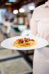 Waiter in white shirt holds plate of spaghetti with tomatoes, herbs, and edible flowers in restaurant setting with blurred background.