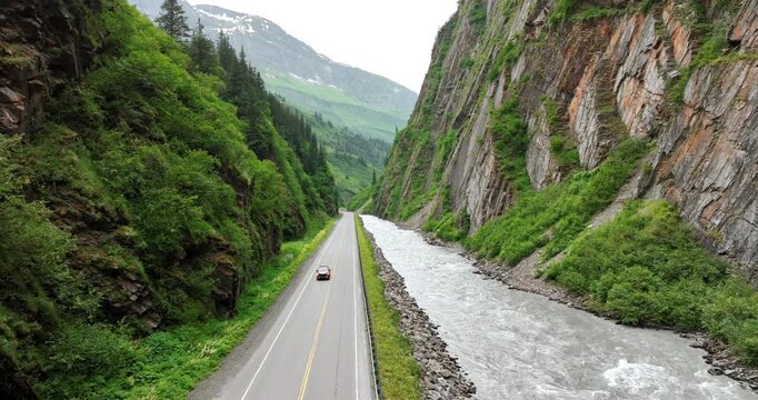 A car approaches by the road built along the fast narrow river at the mountain foot. Travel by the gorgeous Alaska.