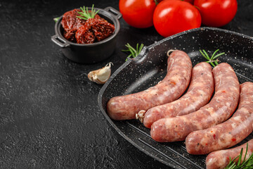 Raw pork sausages seasoned with herbs and spices arranged in a grill pan with garlic, tomatoes, and sun-dried tomatoes on a black textured background. copy space, top view.