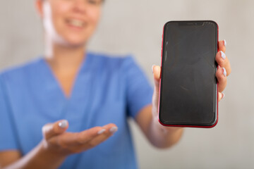 Female nurse in white coat extending hand with phone and showing screen, selective focus on gadget....
