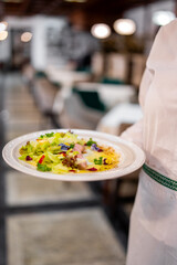 Professional waiter in white uniform presenting a gourmet salad with greens, edible flowers, and meat slices in an upscale restaurant. Blurred background enhances focus.