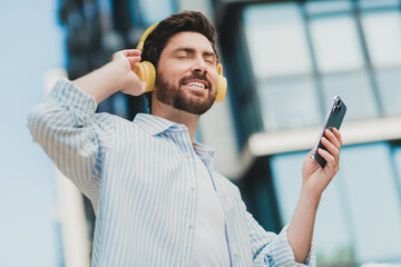 Young man enjoys music with headphones while holding a smartphone in a modern urban area on a sunny...