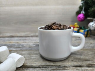 white cup filled with coffee beans next to pieces of marshmallows. There is a Christmas tree in the background