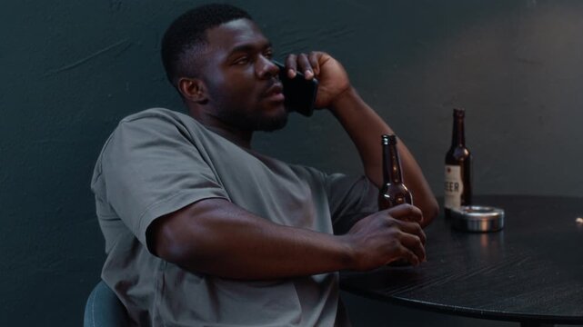 Tilt up shot of young Black man under house arrest wearing electronic ankle monitor sitting at table drinking beers and talking on phone