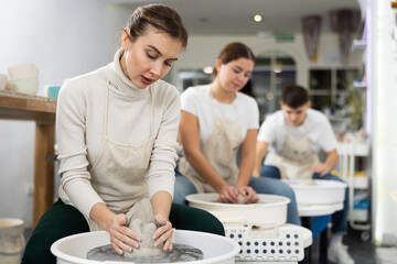 Positive young woman in apron working with pottery wheel in workshop. Persons engaged in pottery
