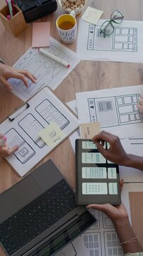 Vertical top view shot of diverse creative team of UI designers developing mobile app interface using digital tablet and analyzing wireframes on cluttered with papers desktop at office, slow motion