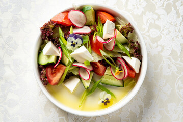 Fresh vegetable salad with tomatoes, cucumbers, radishes, lettuce, green onions, cheese cubes, and edible flowers in a white bowl on patterned tablecloth, top view.