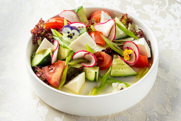 Fresh vegetable salad with tomatoes, cucumbers, radishes, lettuce, green onions, cheese cubes, and edible flowers in a white bowl on patterned tablecloth, top view.