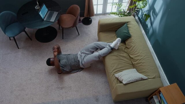 Zoom out shot of sporty Black man under house arrest wearing electronic monitoring bracelet lying on floor with legs on couch and doing crunches, training his abs and strengthening core muscles
