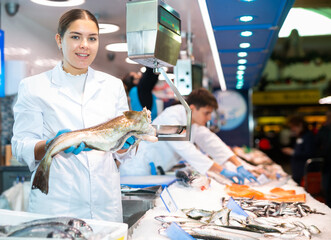 Positive young saleswoman demonstrating cod fish behind counter in fish store