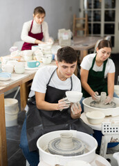 Smiling young man in apron holding handmade clay bawl sitting at pottery wheel in workshop. Persons working with potter wheel