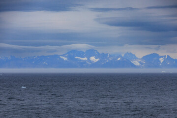 View from a cruise ship toward Greenland’s raw Arctic landscape with snowy mountains, dark sea, and dramatic cloudy sky.