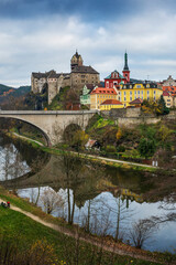 View of Loket Castle and colorful historic buildings in Loket, Czech Republic, with a stone bridge and river reflection on a cloudy autumn day