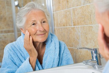 Woman in blue bathrobe looks at her reflection in the mirror, feeling happy and confident. Daily routine for elderly.