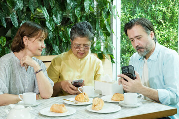 Asian senior woman and two Caucasian friends sit at a cafe table, smiling as they use smartphones and enjoy pastries and tea, sharing a joyful and relaxed retirement moment together.