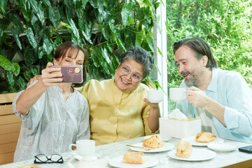 Asian senior woman and two Caucasian friends smile and raise teacups while taking a cheerful selfie at a cafe table, enjoying pastries and sharing a joyful retirement moment together.