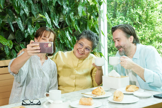 Asian senior woman and two Caucasian friends smile and raise teacups while taking a cheerful selfie at a cafe table, enjoying pastries and sharing a joyful retirement moment together.