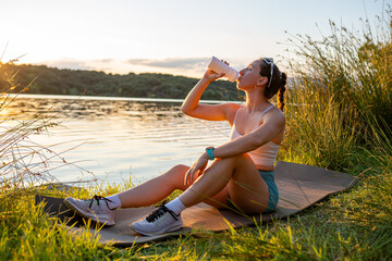 Young athlete drinking water after training by the lake at sunset