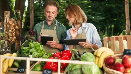 Caucasian happy pretty family couple working selling food at market at counter. Portrait. Man writing in notepad while woman typing on tablet checking products doing inventory. Business, healthy food