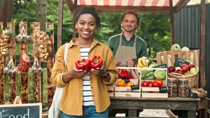 African American woman outdoor buying food at food market. Woman buy vegetables and paying with smartphone pay pass. Male and female family of farmers work at market selling organic products