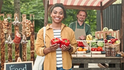 African American woman outdoor buying food at food market. Woman buy vegetables and paying with smartphone pay pass. Male and female family of farmers work at market selling organic products