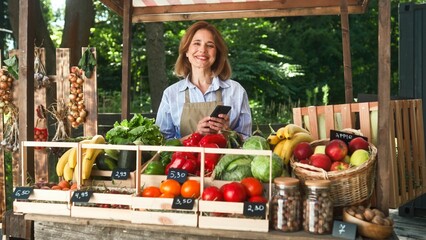 Cheerful mature woman farmer typing on mobile phone and smiling to camera standing at counter selling fresh vegetables and fruits. Local food market. Happy adult saleswoman working at market