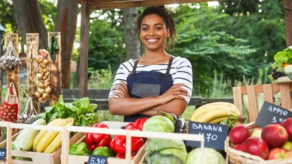Camera moving to pretty young smiling African American woman farmer in apron selling farm food at market outdoor. Beautiful positive happy female sell organic fresh vegetables and fruits