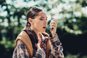 Young woman outdoors using an inhaler during an asthma episode on a bright sunny day in a green park