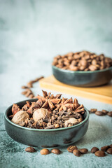 Close-up bowl filled with star anise, nutmeg and cloves in front of blurred coffee beans, captured in soft daylight on textured grey stone for fragrant autumn beverage concepts