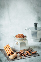 Moka-pot basket brimming with ground coffee among scattered beans, cinnamon sticks, nutmeg, star anise and cloves, with blurred aluminium espresso maker in the back
