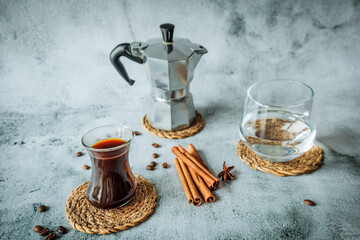 Black coffee in curved glass with water tumbler, moka pot and cinnamon sticks on woven coasters, scattered beans on pale stone background, airy vertical layout for cafe ads