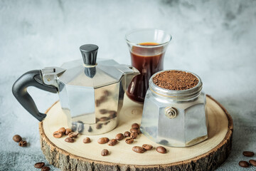 Stovetop moka pot, basket packed with ground coffee and freshly brewed cup arranged on a rustic wood slice, scattered beans over light concrete, cozy cafe vibe